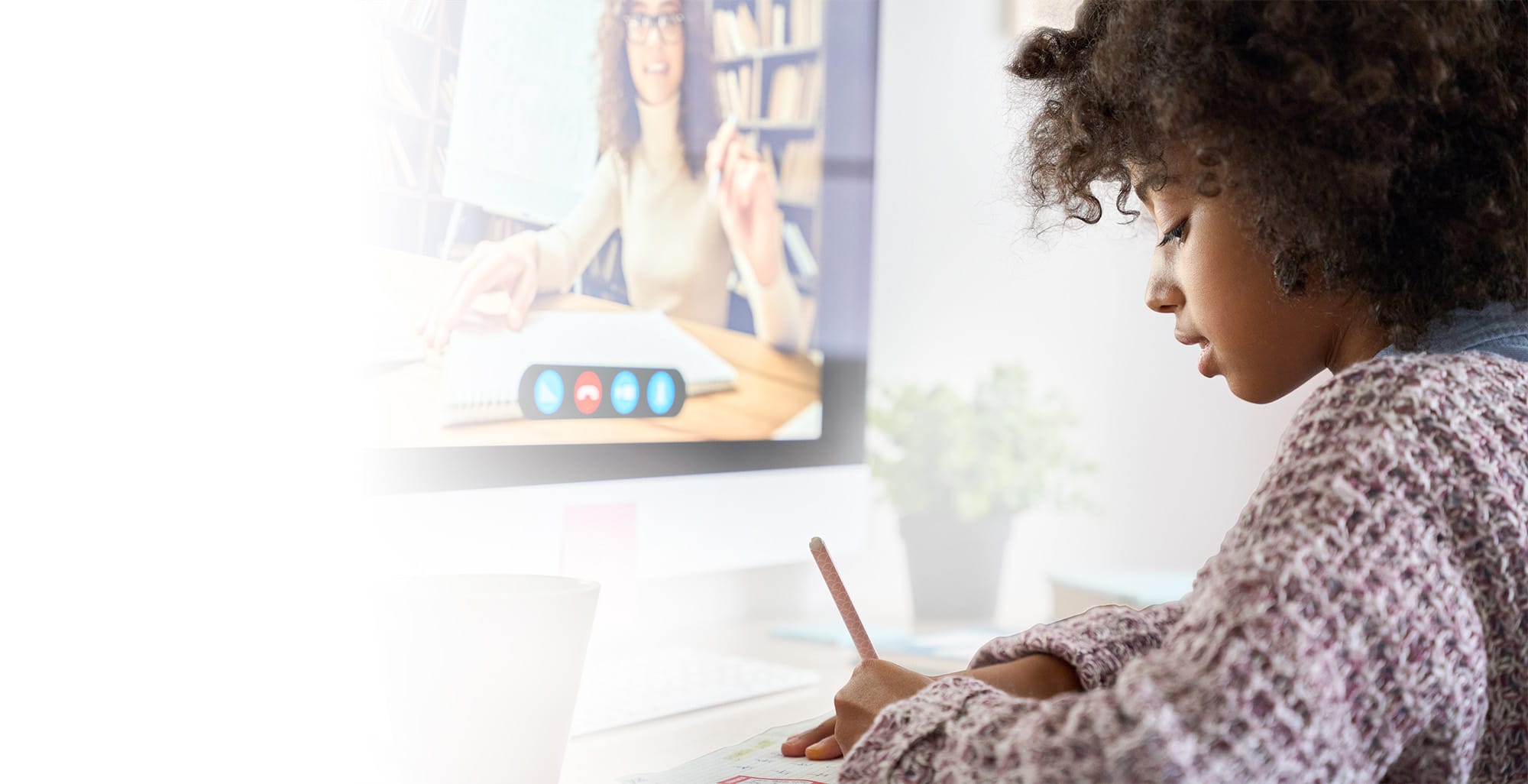 A young woman writes in a notebook while attentively watching an online lesson on her computer, where a teacher is visible on the screen during a video call.