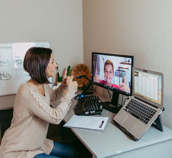 A woman sits at a desk, smiling and giving a thumbs up during a dyslexia therapy video call with a boy on her monitor, while a laptop and papers are open in front of her.