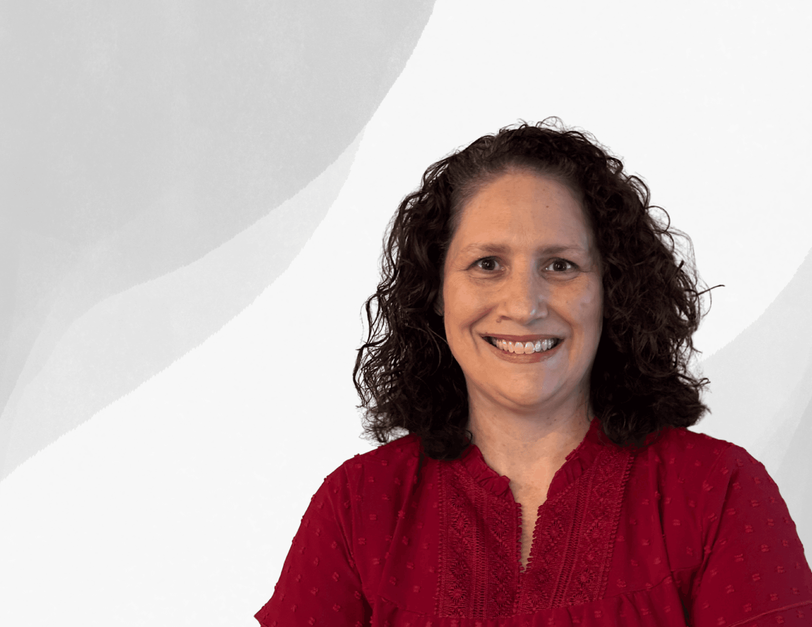 A woman with curly brown hair, wearing a red blouse, smiles while standing in front of a white and gray abstract background.