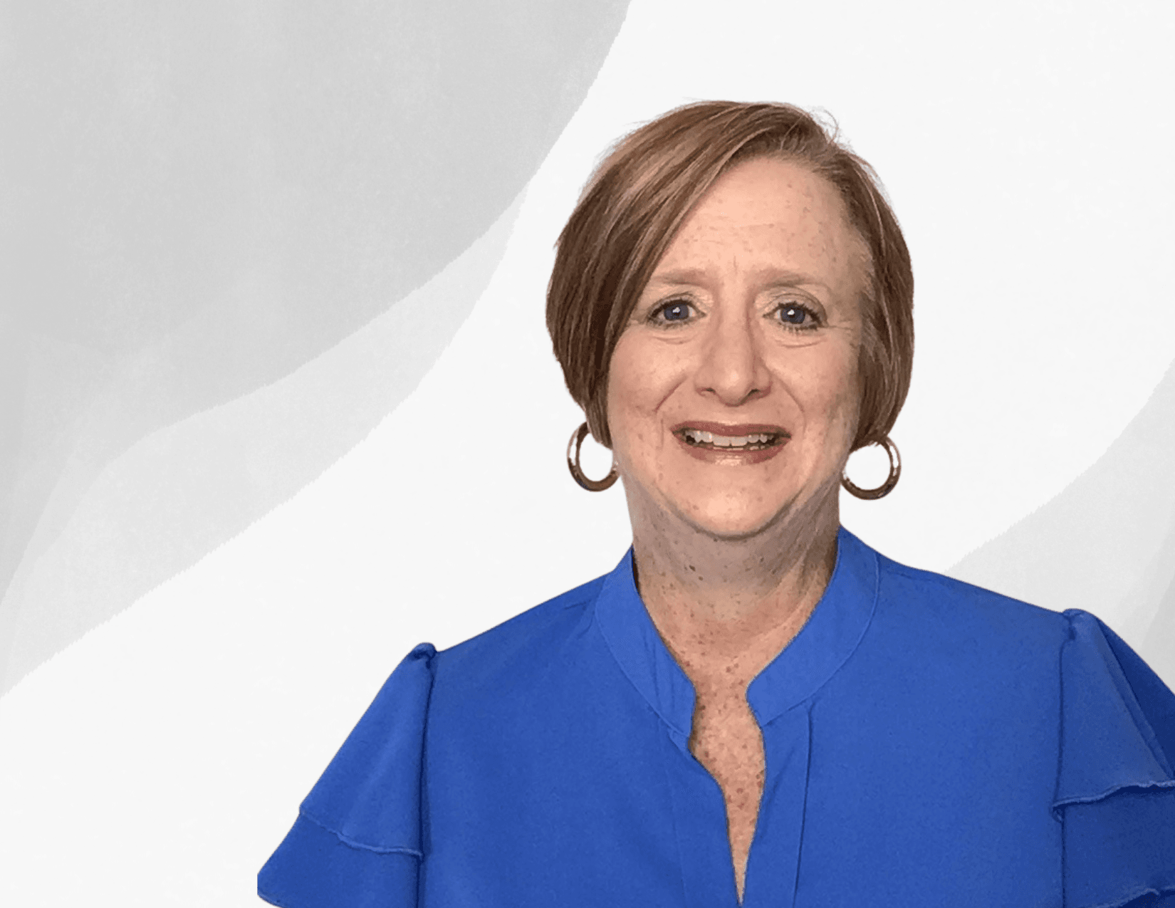 A woman with short light brown hair, wearing hoop earrings and a royal blue blouse, smiles in front of a light gray and white abstract background.