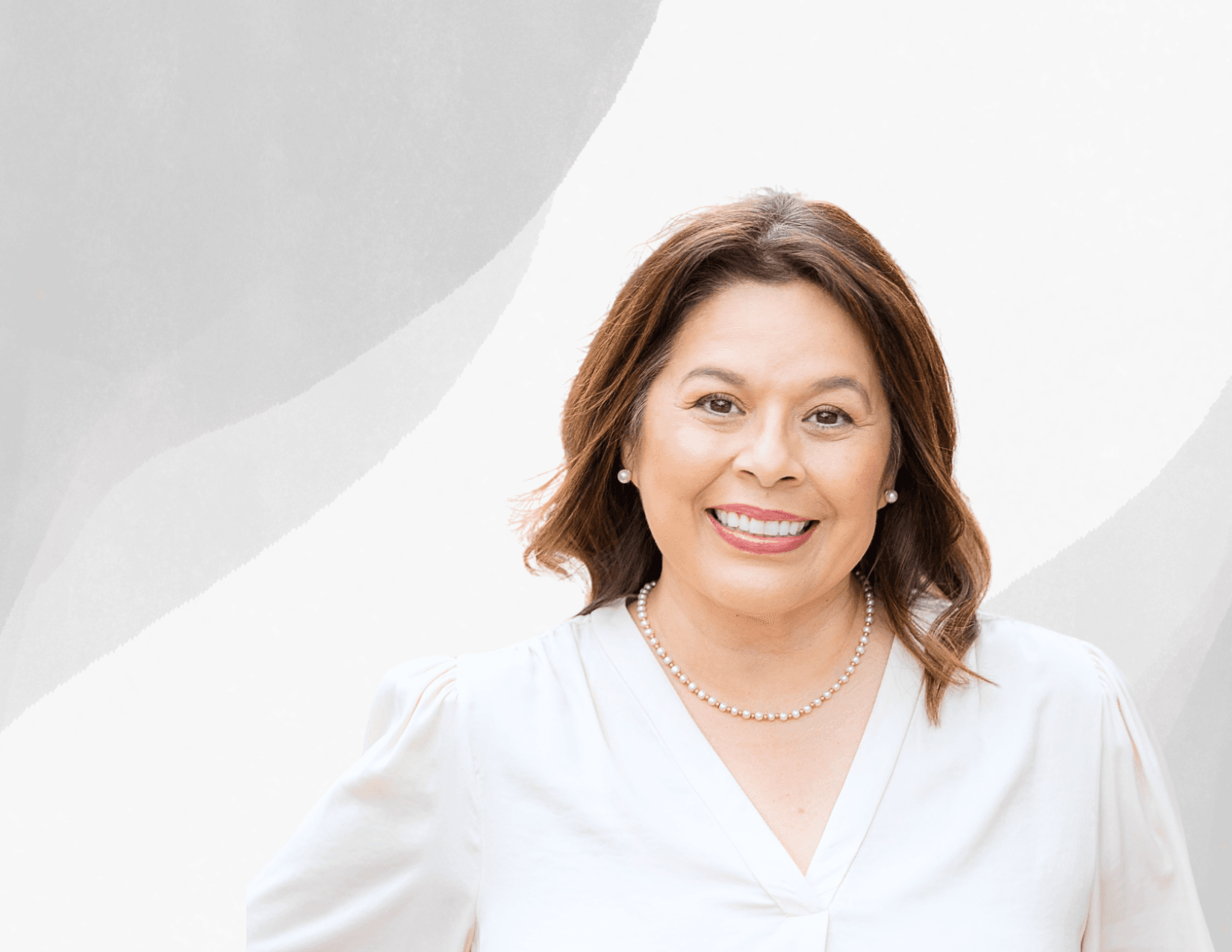 A woman with medium-length brown hair, wearing a white blouse and pearl necklace, smiles at the camera against a light gray abstract background.