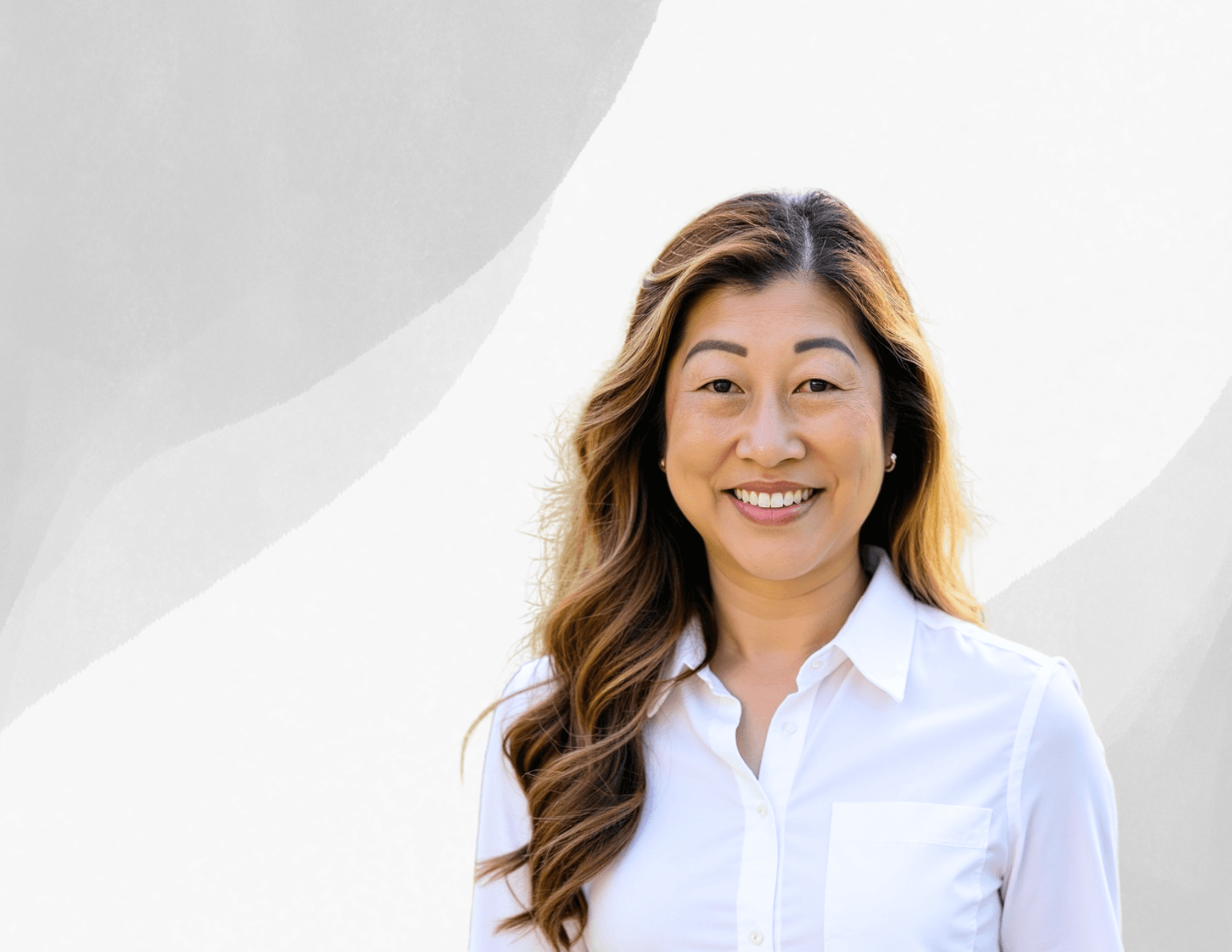 A woman with long wavy hair wearing a white button-up shirt smiles while standing in front of a light, abstract background.