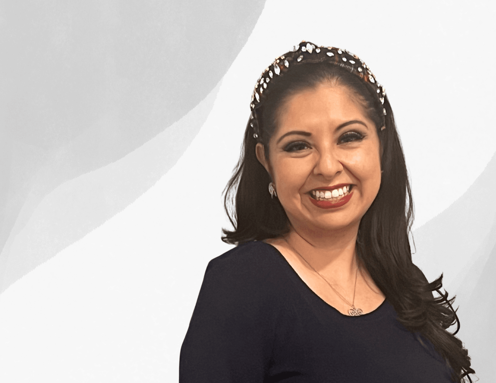 A woman with long dark hair, wearing a dark top and a jeweled headband, smiles at the camera against a light gray and white abstract background.