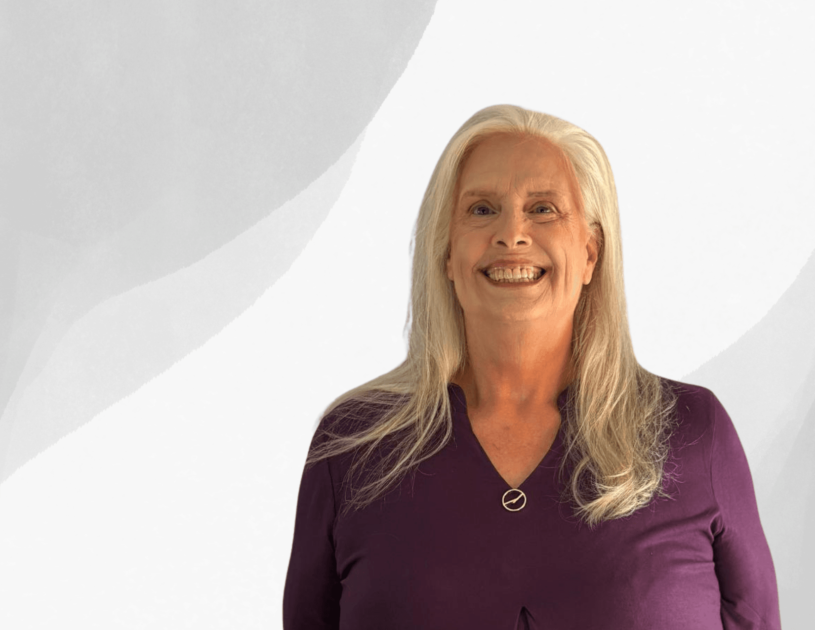 A smiling older woman with long white hair wears a purple top and a necklace, standing against a light gray and white abstract background.