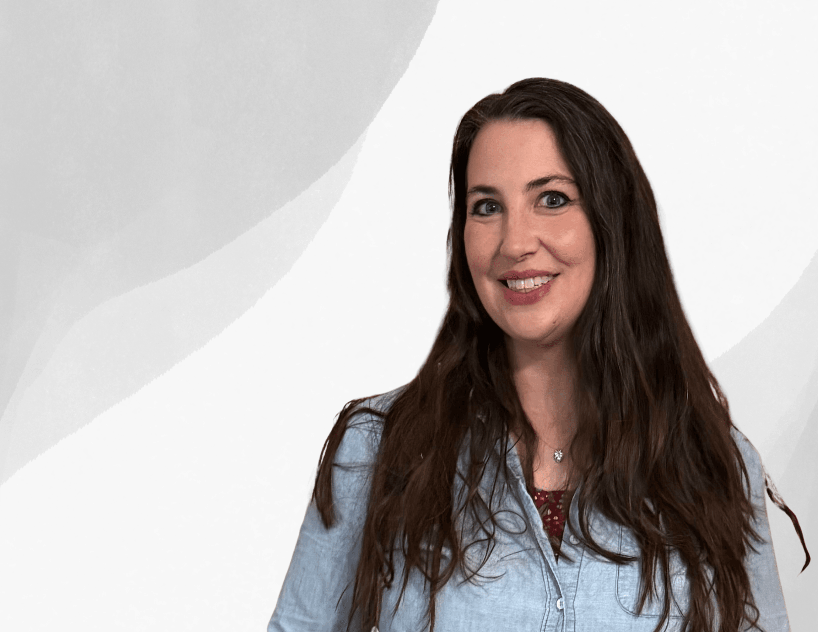A woman with long brown hair wearing a light blue button-up shirt and red top smiles in front of a white and gray abstract background.