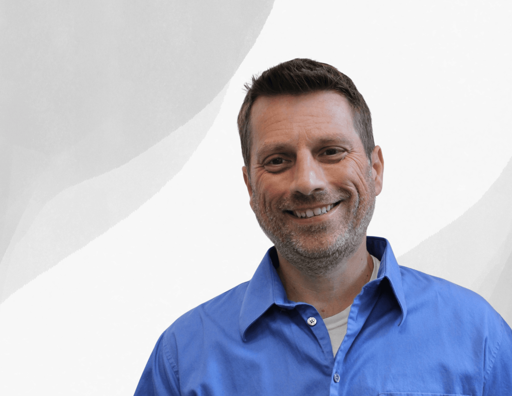 A man with short brown hair and a beard smiles at the camera. He is wearing a blue collared shirt over a light gray t-shirt, standing against a light background with subtle gray curved shapes.