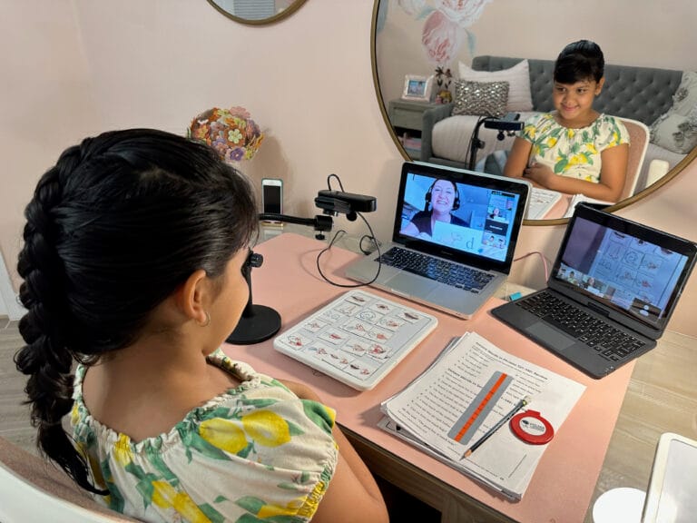 A young girl with a braided ponytail sits at a desk, engaging in an online lesson with a teacher displayed on a laptop. Educational materials and another laptop are on the desk; a mirror reflects her face.