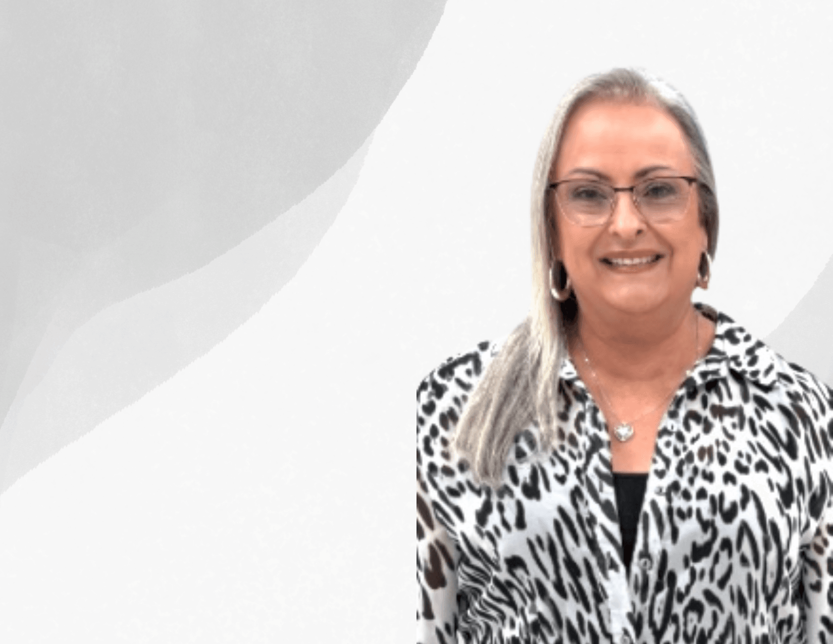A woman with long, straight gray hair and glasses smiles at the camera. She is wearing hoop earrings, a necklace, and a black-and-white animal print blouse, standing against a light gray abstract background.