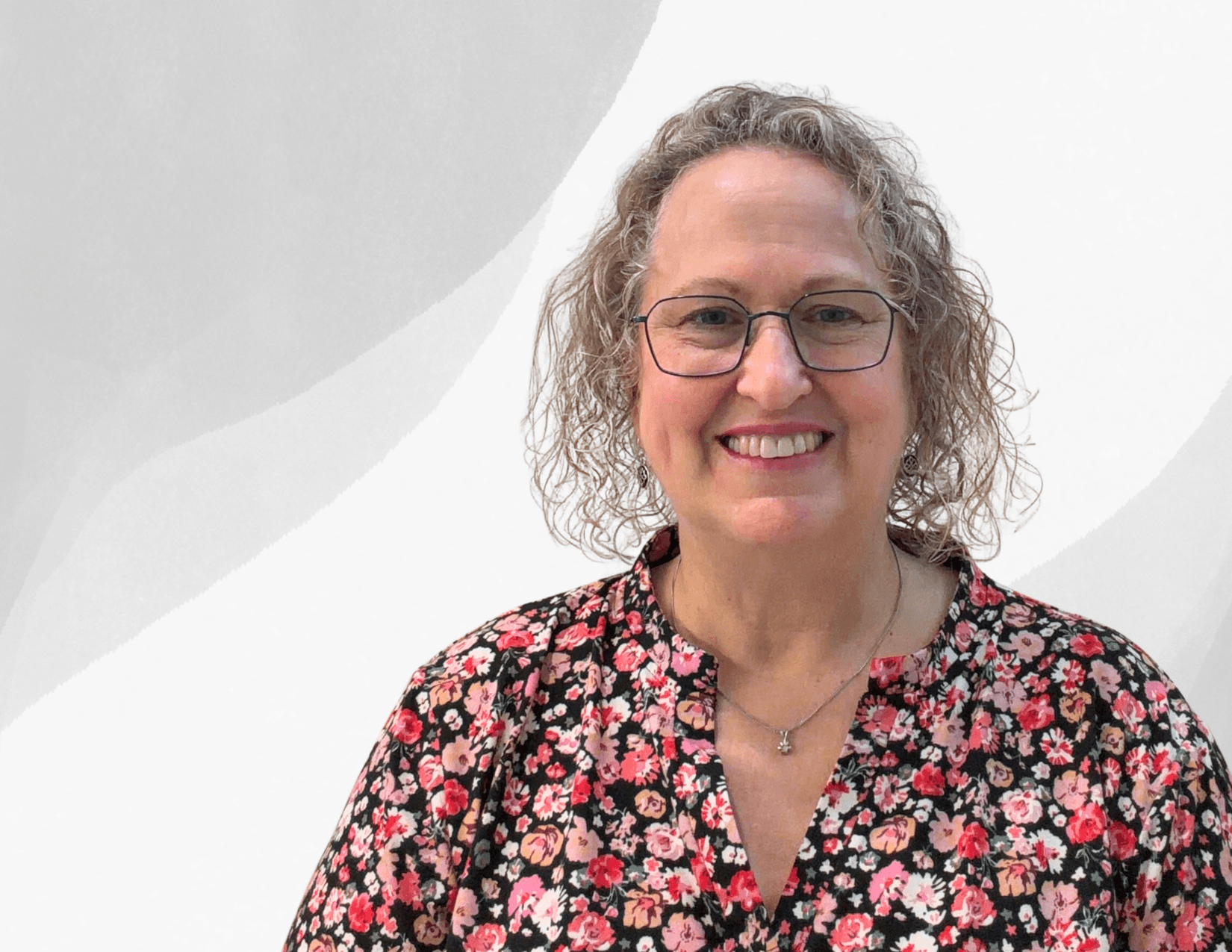 A woman with curly gray hair and glasses smiles at the camera. She is wearing a black top with a pink floral pattern and a necklace, standing against a light, abstract background.